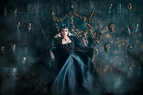 Queen in a black gown sits on a throne surrounded by a wall of masks