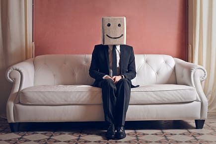 man in a business suit sitting on a sofa with a box on his head; the box has a smiley face drawn on with marker